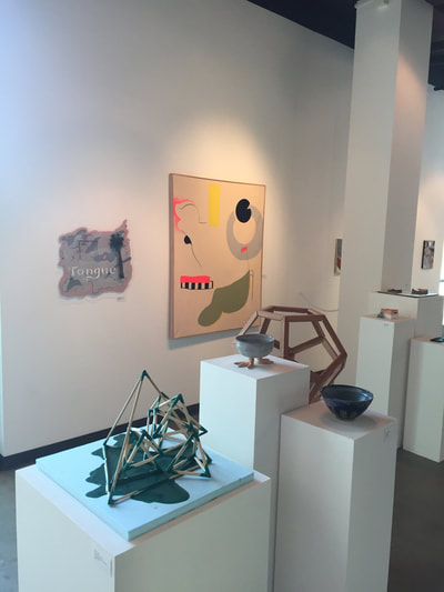 Photo of closeup of three different sculpture object sitting on pedestals. The first object is a spherical pentagon made of wood, the second object is a ceramic bowl, the third object is a structure made of wood dowels. Back of photo: gallery wall lined with paintings.