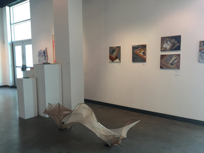 Photo of art pieces hanging on gallery wall. And a sculpture piece made of metal and mesh material.